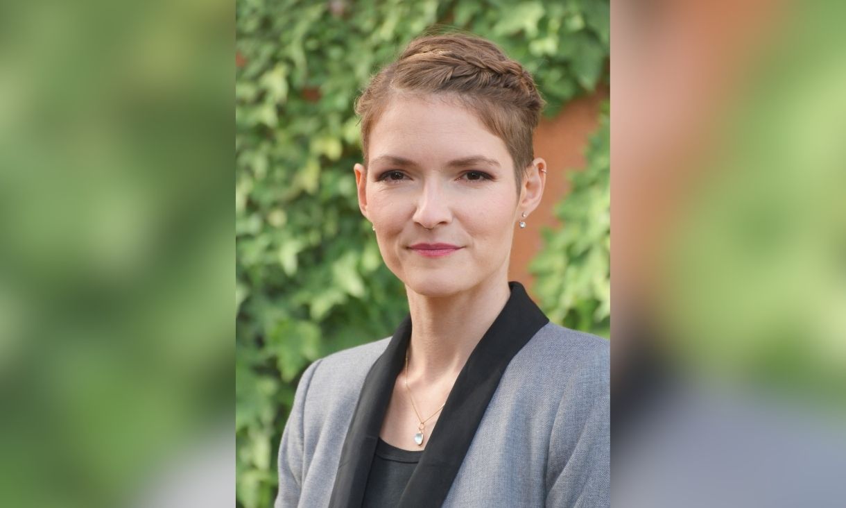 Dr Kate Gerber is pictured in front of a green leafy background, wearing a grey suit jacket. 