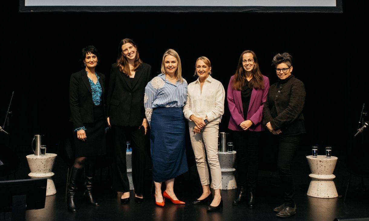 Six women stand on a dark stage with a projector behind them.