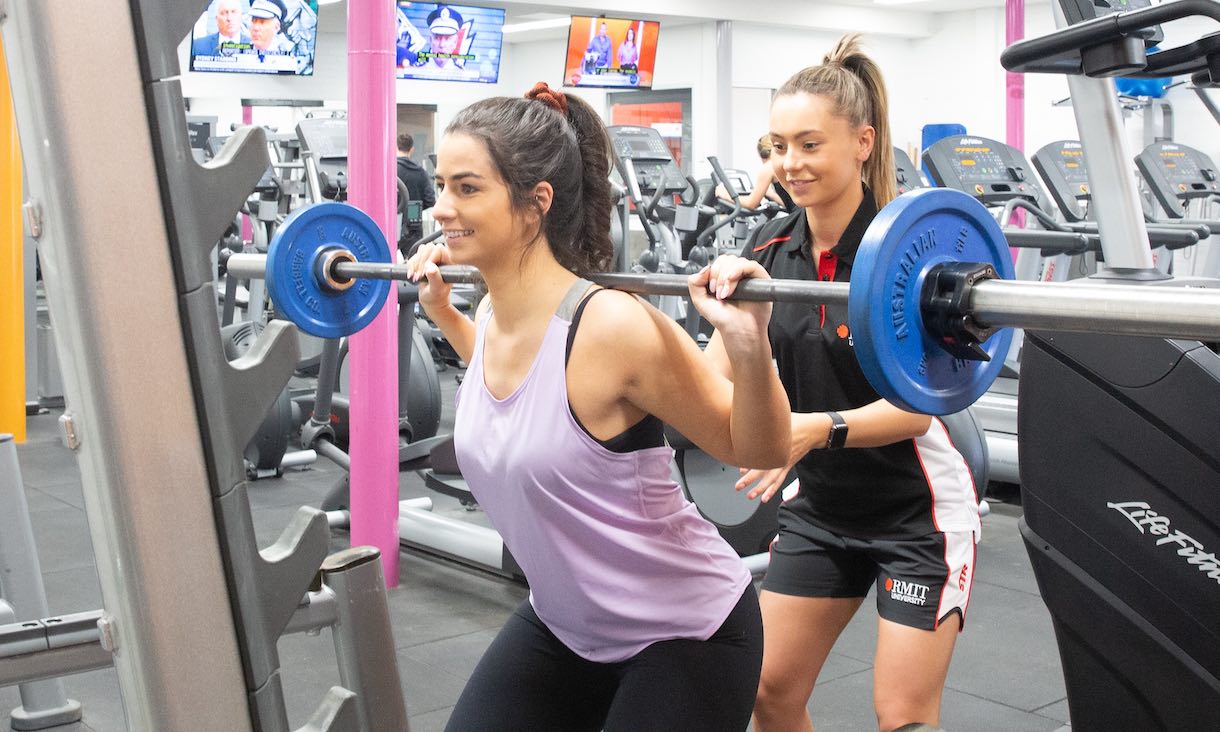 two women in a gym doing weighted squats, one is a personal trainer in rmit gear advising the other